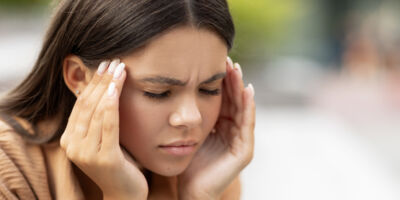 Tired young woman suffering from headache, sitting on bench outdoors at university campus, rubbing her temples, have migraine, feeling exhausted, panorama with copy space, closeup